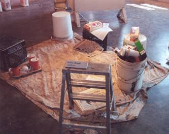 installation of coffee break comprise of donuts and newspapers on overturned milk crates and five-gallon buckets, a step-ladder, and trash: coffee cups, cigarette butts, and empty nips